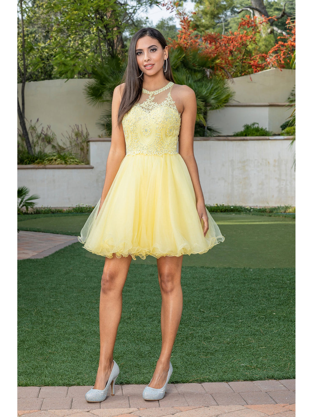 Outdoors, a woman models the Beaded Embroidered Lace Short Babydoll Party Dress in yellow—sleeveless, knee-length with a fitted bodice and flared skirt—styled with silver high heels amid greenery and a white wall.