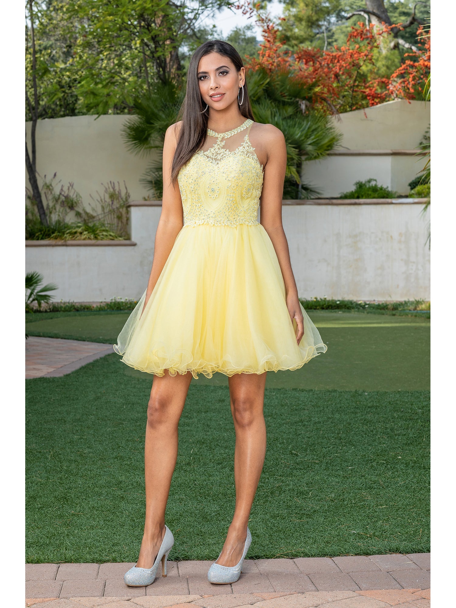 Outdoors, a woman models the Beaded Embroidered Lace Short Babydoll Party Dress in yellow—sleeveless, knee-length with a fitted bodice and flared skirt—styled with silver high heels amid greenery and a white wall.